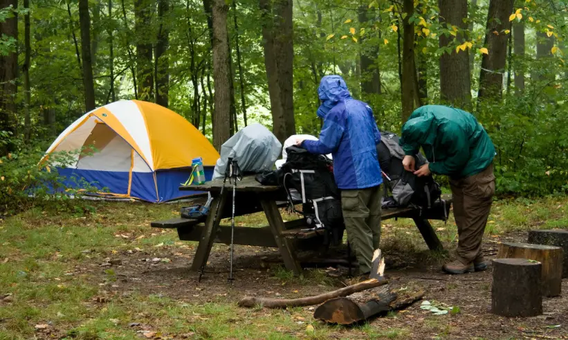Two people opening rucksacks on a park bench with a yellow, blue and white tent nearby.